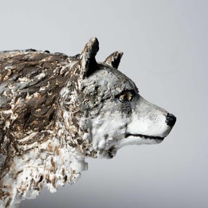 Può includere: Scultura dettagliata della testa di un lupo, con una superficie strutturata in tonalità di grigio, marrone e bianco. Il profilo del lupo è visibile, con un focus sulla sua espressione attenta e sui dettagli. Una rappresentazione artistica della fauna selvatica.