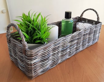 May include: A rectangular, woven gray wicker basket with two arched handles. Inside the basket sits a small potted artificial green plant, a white cylindrical candle, a clear bottle with a black cap containing green liquid, and a clear container of cotton swabs.