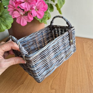 Könnte beinhalten: Ein rechteckiger Weidenkorb mit zwei gebogenen Griffen. Der Korb ist in Blau-, Grau- und Brauntönen bemalt. Ein brauner Lederriemen ist mit einer Messingniete an der Seite befestigt. Eine Hand hält den Korb, mit einer rosa blühenden Pflanze im Hintergrund.