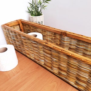 May include: A woven wicker basket with a wooden frame, designed to hold toilet paper rolls. The basket is filled with two rolls of toilet paper and is sitting on a wooden surface.