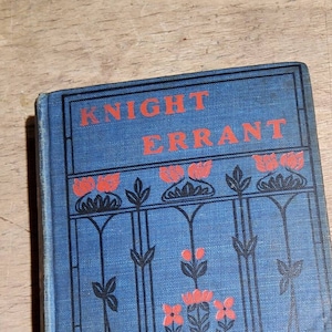 May include: A vintage book titled "KNIGHT ERRANT" with a blue cover. The cover features red text and floral designs in black and red. The book has a rectangular shape and is placed on a wooden surface.