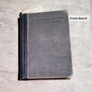 May include: An antique book with a dark gray cover, titled "Cooley's Series" and "Physical Science." A small newspaper clipping is tucked inside. The book shows signs of wear, suggesting age and use.