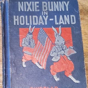 May include: Vintage children's book titled "NIXIE BUNNY IN HOLIDAY-LAND" with illustrations of anthropomorphic rabbits. The cover features rabbits in suits and red pants, holding an American flag. The book has a blue cover with the author's name, SINDELAR.