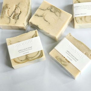 May include: Four bars of handmade soap with a white label that reads "Immortelle Soap Company" and "Apple Sage Soap" with the text "nourishing & moisturizing" and "5oz (140g)". The soap is a light beige color with green swirls.