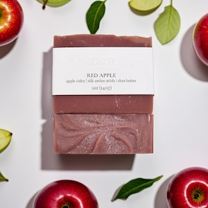 May include: Two bars of red apple soap with a white label that reads "RED APPLE" and lists ingredients. The soap is surrounded by red apples, green leaves, and apple slices. The soap weighs 5 oz (140g).