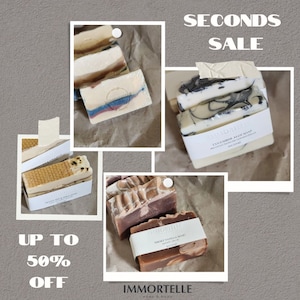 May include: A collection of rectangular soap bars labeled Immortelle, including Cucumber Aloe Soap with black swirls, Honey Bee & Milk Soap with a honeycomb texture, and Smoky Vanilla Soap. The image features text reading SECONDS SALE and UP TO 50% OFF.