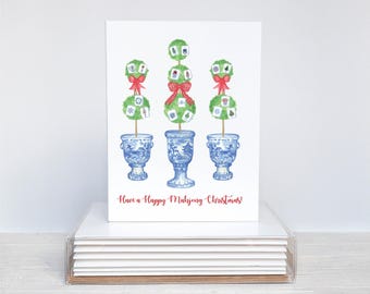 May include: A watercolor Christmas card featuring three topiary trees with red bows, set in blue and white porcelain vases. The card reads "Have a Happy Mahjong Christmas!" in red script, with a stack of cards in a clear acrylic box.