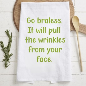 May include: White kitchen towel with green text that reads "Go braless. It will pull the wrinkles from your face." The towel is on a wooden cutting board with a wooden spoon. Rosemary sprigs are on the left.