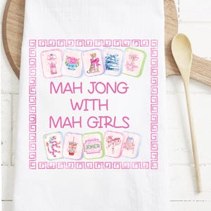 May include: White kitchen towel with a pink border and the text "MAH JONG WITH MAH GIRLS". The towel features watercolor illustrations of Mahjong tiles. A wooden spoon rests on a wooden cutting board in the background.