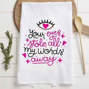 May include: White kitchen towel with a pink and black design. The design features a stylized eye with a heart in the center and the text "Your eyes stole all my words away".