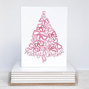 May include: A white Christmas card with a pink bow-shaped tree design. The words "Merry Christmas" are written in cursive below the tree. The card is presented in a clear acrylic holder.