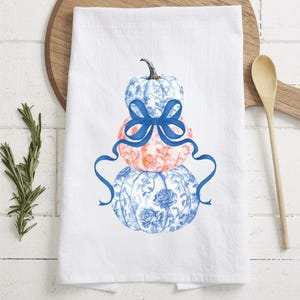 Op de afbeelding: Witte keukendoek met een ontwerp van drie gestapelde pompoenen. De bovenste en onderste pompoenen hebben een blauw bloemenpatroon, de middelste is oranje. Een blauw lint siert de pompoenen. Een houten lepel en plank staan op de achtergrond.