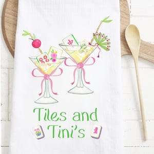 May include: White kitchen towel featuring two martini glasses with Mahjong tiles, pink bows, and decorative garnishes. The towel has the text "Tiles and Tini's" in green. Decorative Mahjong tiles are also shown.