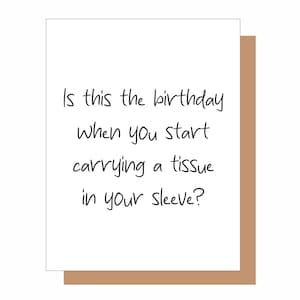 May include: A white rectangular greeting card with black handwritten text that reads: Is this the birthday when you start carrying a tissue in your sleeve? The card is positioned against a solid tan background, showing a clean and simple design.