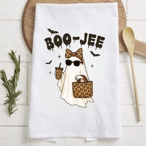 May include: White kitchen towel with a cartoon ghost wearing sunglasses and a leopard print bow, holding a coffee and a tote bag. The towel has the text "BOO-JEE" and bat illustrations.