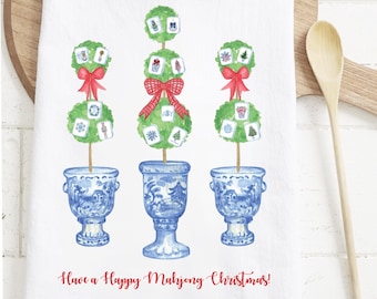 Op de afbeelding: Witte keukendoek met een aquarelontwerp van drie topiary bomen met rode strikken en blauw-witte vazen. Elke boom heeft Mahjong tegels. De tekst "Have a Happy Mahjong Christmas!" is in het rood geschreven.