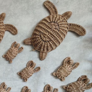 Puede incluir: Una colección de decoraciones de pared con forma de tortugas marinas hechas de mimbre trenzado de color marrón claro en varios tamaños. Las tortugas presentan patrones trenzados detallados en sus caparazones y aletas, dispuestos dispersos.