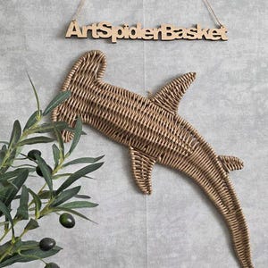 May include: A woven, brown hammerhead shark wall hanging. The shark is made of woven material and is hanging on a gray wall. The sign above the shark reads "ArtSpiderBasket". Olive branches with green leaves and black olives are in the lower left corner.