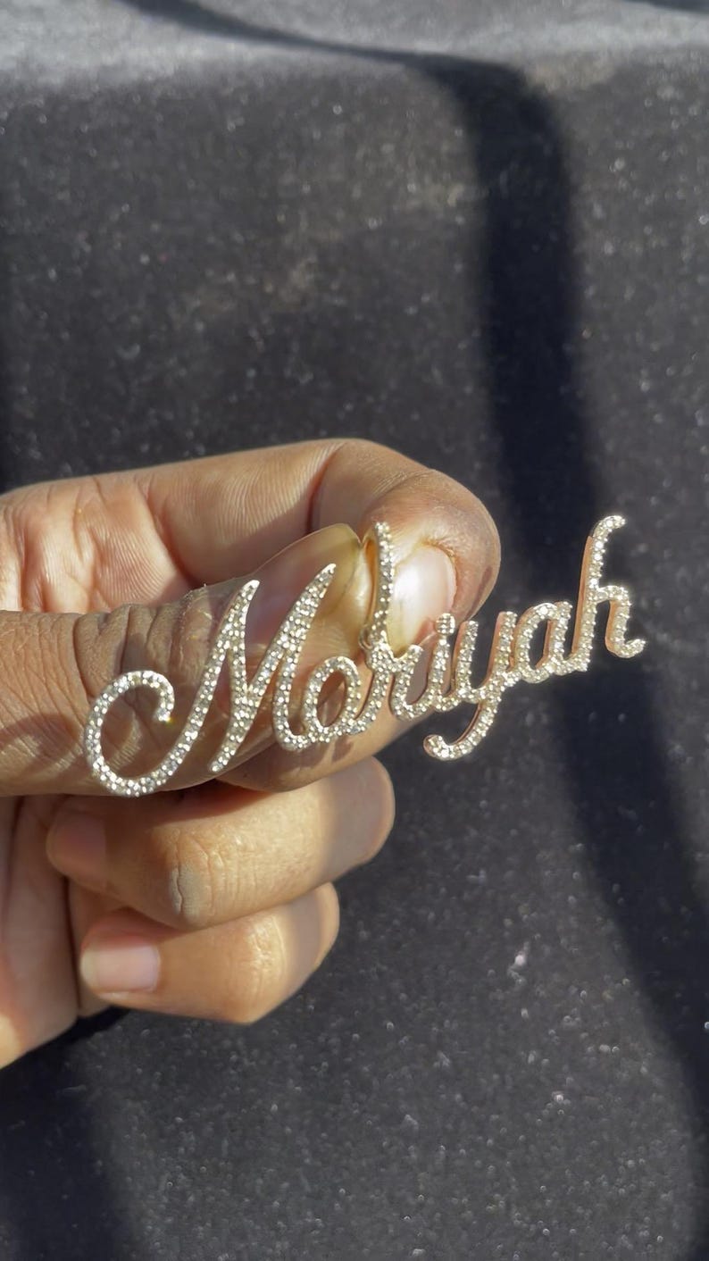 May include: A gold-colored pendant spelling "Mariyah" in a cursive font, encrusted with small, sparkling gemstones. The pendant is held in a hand against a dark background, highlighting the jewelry's details.