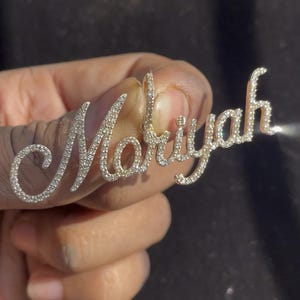 May include: A gold-toned pendant spelling out the name "Mariyah" in a cursive script. The letters are encrusted with small, sparkling gemstones, creating a shimmering effect. The pendant is held up against a dark background.