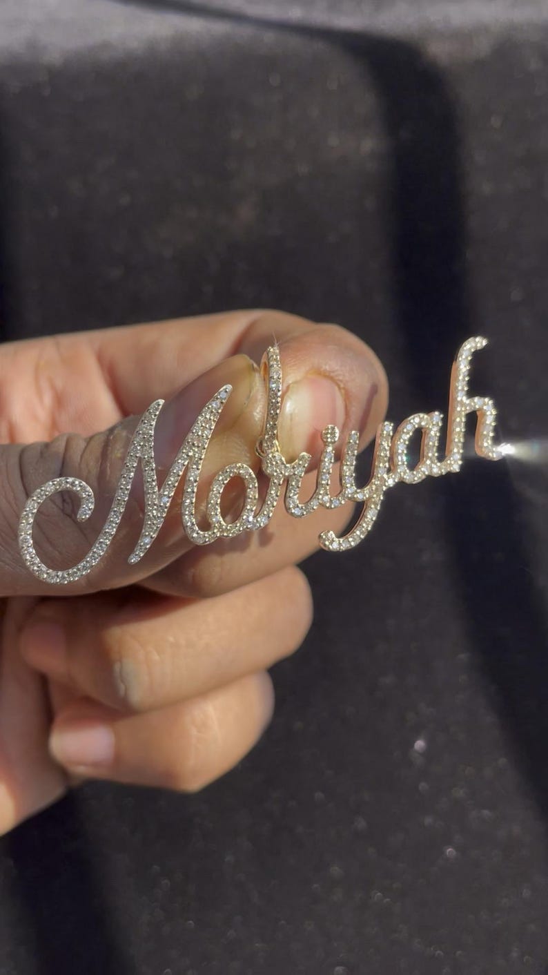 May include: A gold-toned pendant spelling out the name "Mariyah" in a cursive script. The letters are encrusted with small, sparkling gemstones, creating a shimmering effect. The pendant is held up against a dark background.