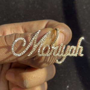 May include: A gold-colored pendant spelling out the name "Moryah" in a cursive font. The letters are encrusted with small, sparkling gemstones. The pendant is held up against a dark background.