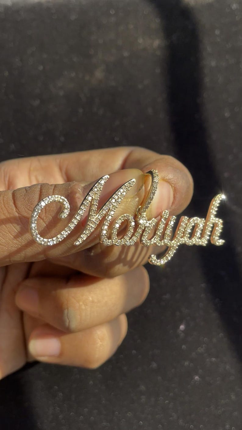 May include: A gold-colored pendant spelling out the name "Moryah" in a cursive font. The letters are encrusted with small, sparkling gemstones. The pendant is held up against a dark background.