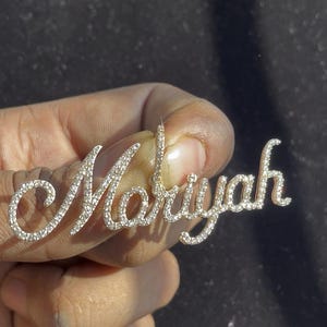 May include: A silver-toned pendant spelling out the name "Mariyah" in a cursive font. The letters are encrusted with small, sparkling crystals. The pendant is held up against a dark background.