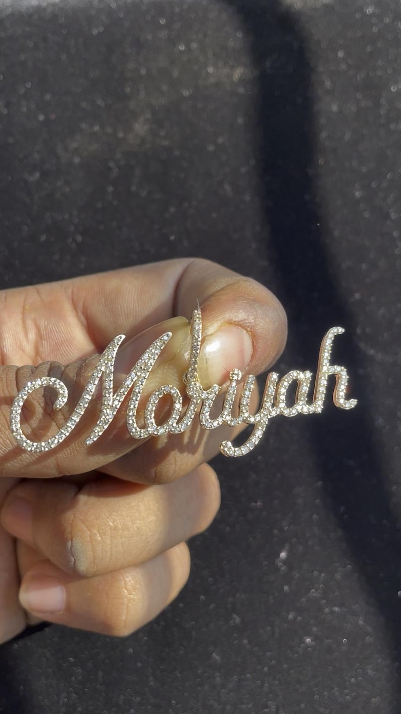 May include: A silver-toned pendant spelling out the name "Mariyah" in a cursive font. The letters are encrusted with small, sparkling crystals. The pendant is held up against a dark background.