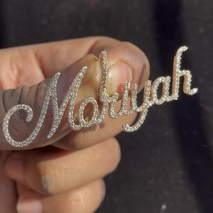 May include: A diamond-encrusted pendant spelling out the name "Mariyah" in a cursive font. The pendant is made of gold and features numerous small, sparkling diamonds. The design is elegant and personalized.