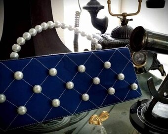 May include: A blue quilted clutch bag with a pearl handle and pearl embellishments. The bag is on a table with a vintage telephone and other decorative items.