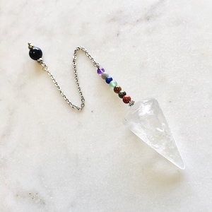 May include: A clear quartz crystal pendulum with a silver chain and colorful chakra beads. The pendulum has a pointed tip and a small, decorative star at the top. The chain connects to a black bead and a silver bead.