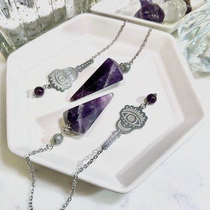 May include: Two amethyst crystal pendulums with silver chains and eye of providence charms. The pendulums are on a white plate.