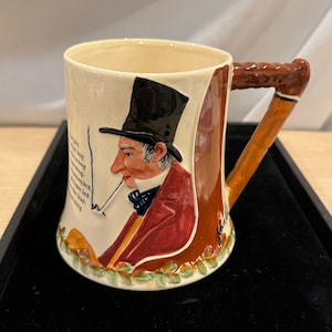May include: Ceramic mug with a detailed illustration of a man in a top hat smoking a pipe. The mug features a brown handle and a cream-colored body with text. The man is wearing a red coat and a black hat.