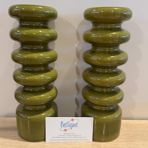 May include: Two olive green ceramic candle holders with a stacked, rounded design. The candle holders have a glossy finish and are displayed on a light-colored wooden surface. A business card with the text "FunTiques" is visible.