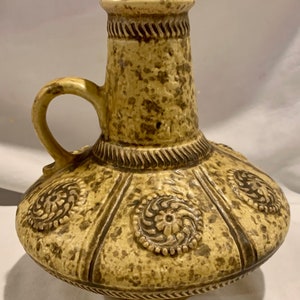 May include: A ceramic vase with a speckled brown and yellow glaze. The vase has a wide base and a narrow neck with a handle. It features a decorative design of brown circles with raised floral patterns.