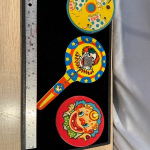 May include: Three vintage tin toys featuring clowns. One toy is a spinning top with a clown in a blue and white polka dot suit. Another toy is a spinning top with a clown in a red and white polka dot suit. The third toy is a clown with a red and yellow hat and a blue and white polka dot suit.