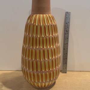 May include: A tall, decorative vase with a textured, vertical pattern in shades of orange and yellow. The vase has a cylindrical neck and a wider, rounded body. A ruler is placed next to the vase for scale.
