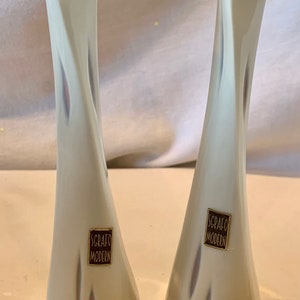May include: Two white ceramic vases with a triangular opening and a swirl design. The vases are decorated with a brown label that reads "Scrafio Modern".