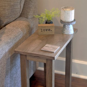May include: A wooden side table with a rectangular top and four legs. The table is topped with a small wooden block that reads "love." A candle in a metal holder and a small coaster are also on the table. A gray sofa is in the background.