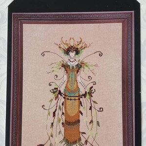 May include: Cross stitch pattern featuring a woman in a brown dress with a leafy design. The pattern is framed in a black and brown frame. The text "Design by Nora Corbett for Wichtelt Imports, Inc." is at the top of the image.