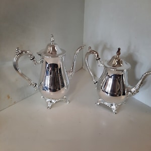 Könnte beinhalten: Zwei silberne Teekannen mit verzierten Griffen und Ausgüssen. Die Teekannen haben dekorative Deckel und stehen auf kleinen, detaillierten Füßen. Die reflektierenden Oberflächen der Teekannen erzeugen ein helles, elegantes Aussehen.