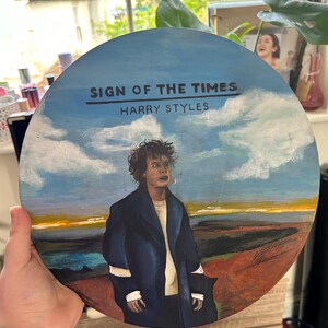 May include: A circular vinyl record painted with a portrait of Harry Styles in a dark coat against a landscape of blue sky, white clouds, and a brown field. The text "SIGN OF THE TIMES" and "HARRY STYLES" is printed in black at the top center.