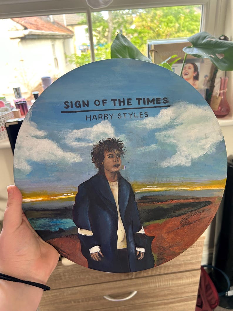 May include: A circular vinyl record painted with a portrait of Harry Styles in a dark coat against a landscape of blue sky, white clouds, and a brown field. The text "SIGN OF THE TIMES" and "HARRY STYLES" is printed in black at the top center.