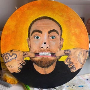 May include: A circular painted vinyl record featuring a portrait of a man with a beard, green eyes, and tattoos on his hands, which are pulling at the corners of his mouth. The background is a vibrant yellow and orange gradient circle.