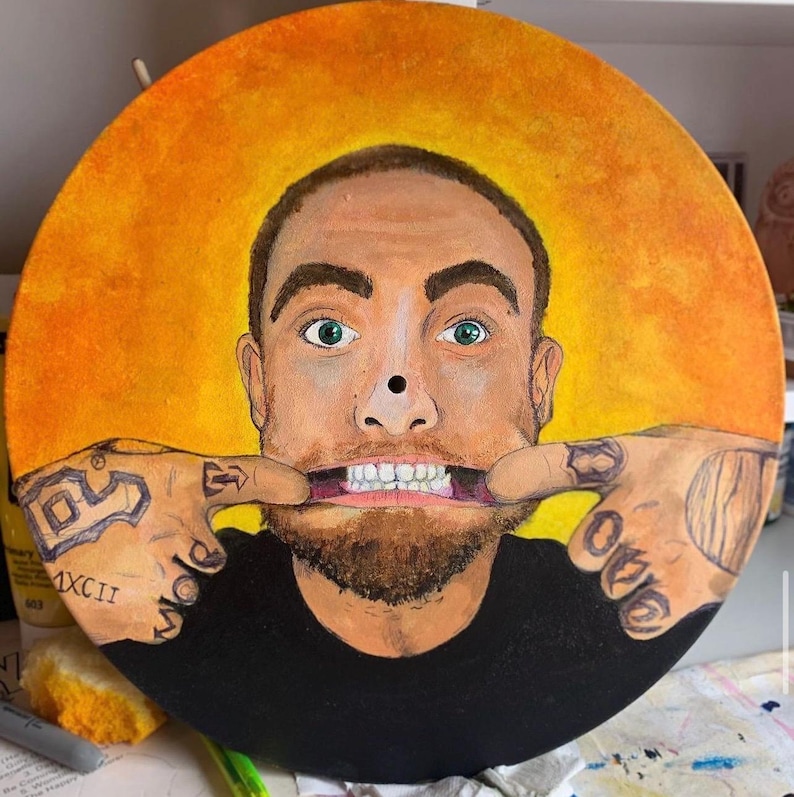 May include: A circular painted vinyl record featuring a portrait of a man with a beard, green eyes, and tattoos on his hands, which are pulling at the corners of his mouth. The background is a vibrant yellow and orange gradient circle.