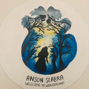 May include: A circular vinyl record painted with a blue and yellow forest scene featuring a silhouette of a person standing among dark trees. The text ANSON SEABRA and WELCOME TO WONDERLAND is printed in black at the bottom of the record surface.