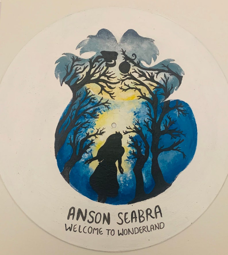May include: A circular vinyl record painted with a blue and yellow forest scene featuring a silhouette of a person standing among dark trees. The text ANSON SEABRA and WELCOME TO WONDERLAND is printed in black at the bottom of the record surface.