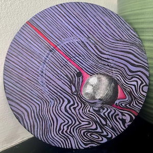May include: A circular vinyl record painted with a purple and black wavy line pattern. A bright red diagonal line cuts across the surface, leading to a central silver sphere that features the text THE TOP printed on its surface.