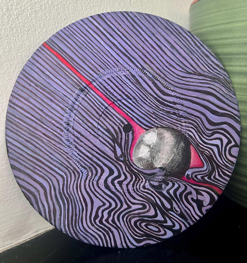 May include: A circular vinyl record painted with a purple and black wavy line pattern. A bright red diagonal line cuts across the surface, leading to a central silver sphere that features the text THE TOP printed on its surface.